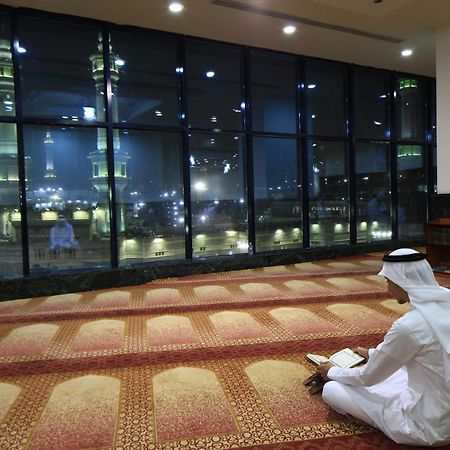 Al Ghufran Safwah Hotel Makkah Mekka Exteriör bild The photo depicts a person sitting on a carpeted floor, facing away from the camera. They are dressed in traditional attire, likely a white thobe and a headscarf. In the background, large windows reveal a nighttime view of a mosque, with illuminated 