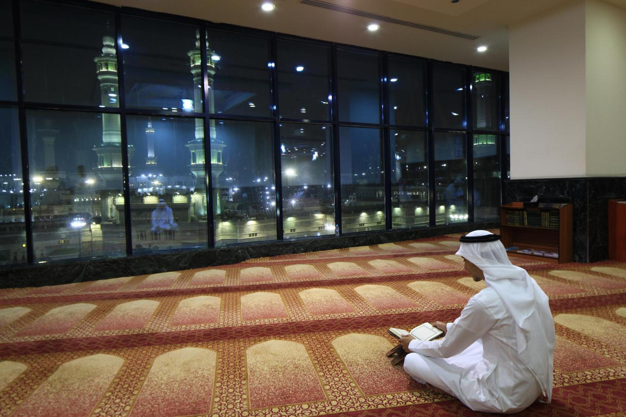 Al Ghufran Safwah Hotel Makkah Mekka Exteriör bild The photo depicts a person sitting on a carpeted floor, facing away from the camera. They are dressed in traditional attire, likely a white thobe and a headscarf. In the background, large windows reveal a nighttime view of a mosque, with illuminated 